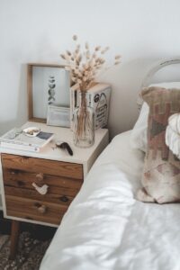 Cozy bedroom interior with wooden white bedside table with glass vase and books bear comfy bed with white sheets against white plain wall