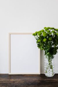 Empty mockup picture in wooden frame and bouquet of green chrysanthemums in stylish glass vase on dark brown wooden table