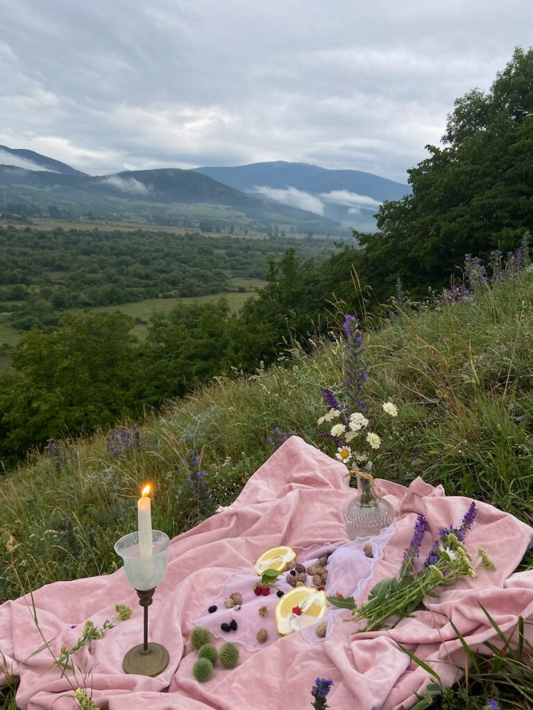 Pink covering with with candles and wildflowers placed on grassy slope of hill in nature with mountains in distance on summer