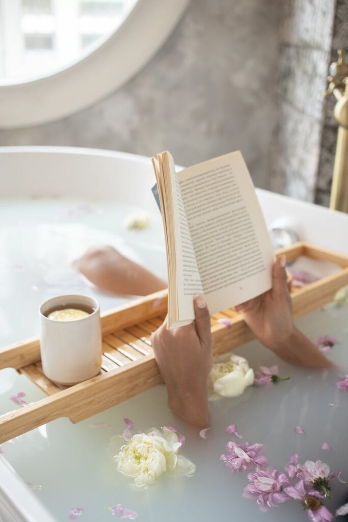 From above of unrecognizable female lying in bathtub full of water with delicate ivory and pink flowers and reading book on blurred background