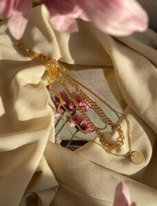 From above of golden chain with pendant placed on crumpled fabric with mirror near pink flowers in light stylish room