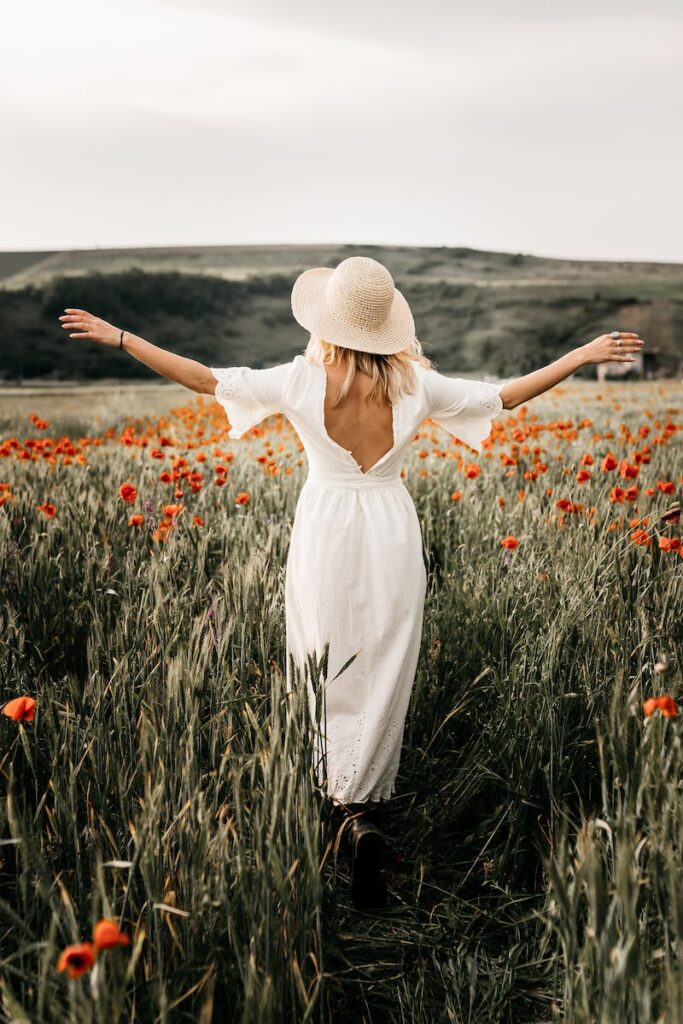 Back view full body of stylish female in white dress and headwear strolling with spread arms on grassy meadow with blooming flowers