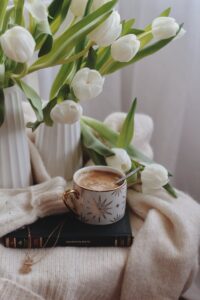 From above of ceramic cup of hot chocolate with spoon placed on book on warm sweater near vases with delicate white tulips